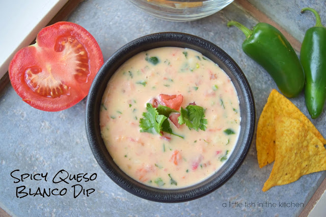 Warm white cheese dip is served in a black bowl. Bits of green peppers, and red tomatoes are visible thoughout the cheese dip. The bowl is garnished with a few whole cilantro leaves and chopped tomato. Two tortilla chips, two whole green jalapeno pepper and a sliced whole tomato surround the bowl for decorative effect.