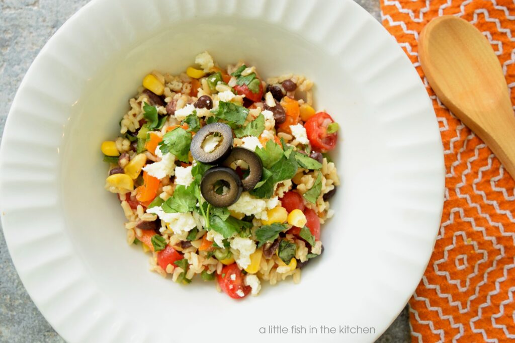 A serving of colorful, fresh Southwestern Brown rice salad is in a white bowl. Bits of fresh red and orange bell pepper, cherry tomato slices, corn kernels, black beans are visible mixed in with fluffy cooked brown rice. The spicy brown rice salad is topped with crumbed cotija cheese, chopped fresh cilantro and slices black olives. 