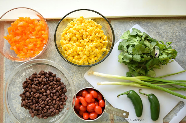 The vibrant ingredients for flavorful Southwestern Brown Rice salad are portioned out: A bunch of fresh cilantro, fresh green onions, two whole, fresh jalapeno peppers, cherry tomatoes, cooked black beans, corn and chopped bell peppers. 