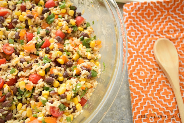 A clear glass serving bowl is filled with freshly prepared, eye-catching Southwest Brown Rice Salad. The colorful dish is chockful of diced fresh peppers, corn kernel, black beans, cherry tomatoes, fluffy brown rice and fresh cilantro and tossed with a zesty honey lime vinaigrette. A orange towel with Southwestern-style stitching lays just next to the serving bowl with a wooded spoon laying on top of the towel.  