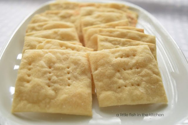 These rustic homemade saltines are lightly golden around the edges, and small bubbly pockets on the tops of the crackers indicate that they are delightfully crisp. The old-fashioned soda crackers are very lightly coated with table salt on the tops and fork prick marks are immediately visible on top each cracker, giving them a familiar appearance. These home saltines are not uniformly cut, as store-bought crackers are, they are comfortingly homemade in their appearance. The crackers are served on a rectagular, white ceramic plate, laid out in two perfect rows. 
