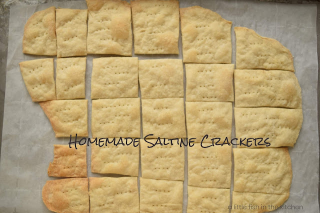 A batch of fresh baked saltine crackers is cooling on a parchment paper-lined baking sheet. The homemade crackers look crisp and lightly toasted on the tops. There are sets of fork prick marks on top of each cracker. The crackers are unevenly cut giving them a rusticly inviting appearance. 