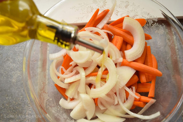 Sliced fresh carrots and onion are drizzled with olive oil in a clear mixing bowl.