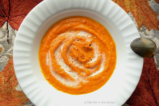 A single serving of vibrantly colored amber soup is in a white ceramic bowl, ready to eat. The soup is eye-catchingly garnishes with small splash of cream and freshly cracked black pepper. A spoon casually rests on the side of the bowl.