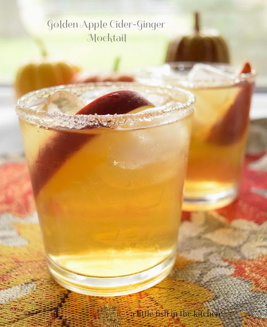A bright golden beverage is served with a cinnamon and sugar rim as well as an apple wedge for garnish. The drinks are festively served on a sparkling silver table runner with orange, red, and gold leaves. 