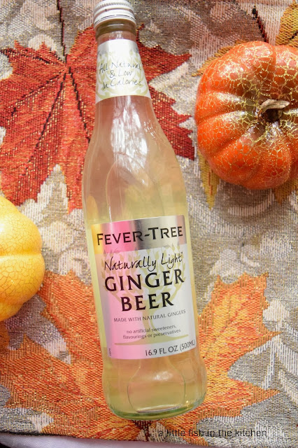 A single bottle of Fever Tree brand ginger beer is laying on a vibrant silver fall table runner. The runner features a pattern of red, gold, and orange autumn leaves. 
