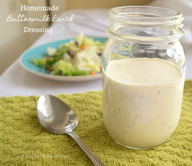 A clear glass mason jar filled with homemade ranch dressing sits on a green cotton tea towel with a spoon on it. A crisp green salad is on a teal plate near the mason jar and the image is slightly blurred in the background. The ranch dressing is white with specks of herbs visible through out the dressing. 