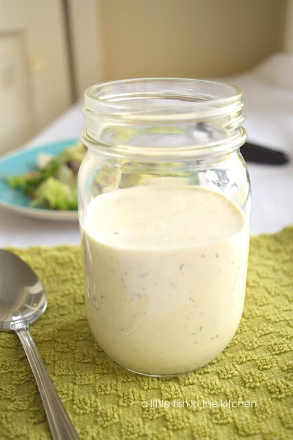 A clear glass mason jar filled with homemade ranch dressing sits on a green cotton tea towel with a spoon on it. A crisp green salad is on a teal plate near the mason jar and the image is slightly blurred in the background. The ranch dressing is white with specks of herbs visible through out the dressing. 