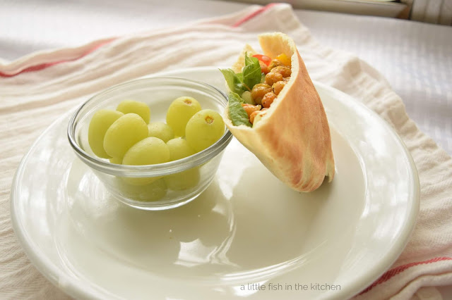 A pita bread stuffed with golden roasted chickpeas, crisp greens looks tempting on a white plate. A small clear ramekin with ripe green grapes is served with the pita sandwich to round out this fabulous meatless meal.