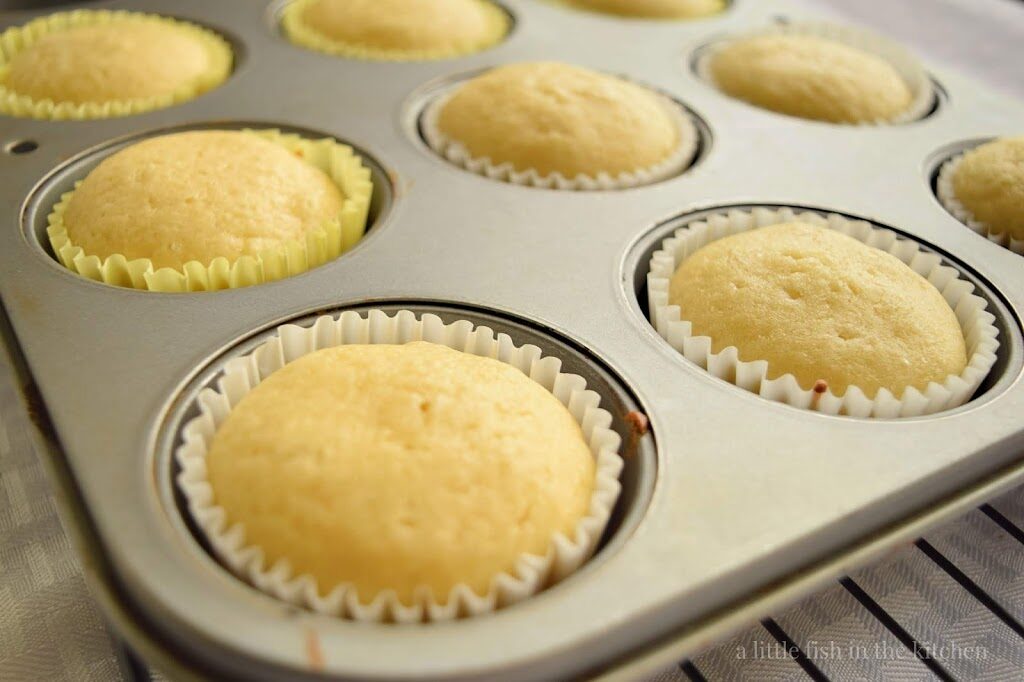 Fresh baked lemon muffins that have been baked into muffins papers are cooling in the baking tin, on a rack. The muffins tops have a light yellow hue to them and they look fluffy and tempting. 