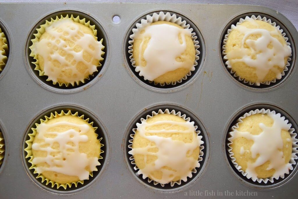 6 fresh-baked, freshly glazed sweet lemon muffins are pictured in the the cavities of a standard metal muffin pan. The muffins tops have a mild yellow tint and a white lemon-flavored glazed has been whimsically drizzled over the tops. 