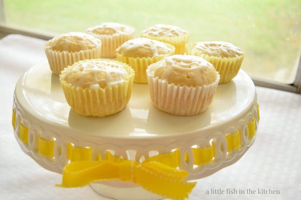 seven fresh lemon muffins are served on a white cake stand that's decorated with a yellow ribbon. The cake stand is on a white table cloth and the table is near a sunny window. The muffins are in yellow and white muffin papers and the the crumb has a light yellow hue. A sweet white glaze is drizzled over the tops of the citrus muffins. 