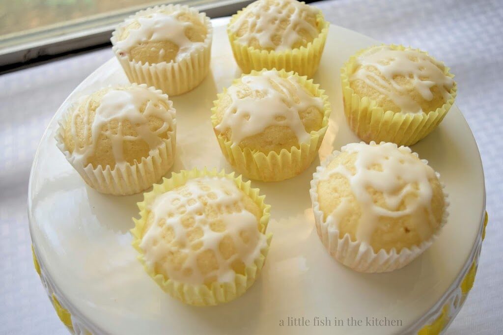 seven fresh lemon muffins are served on a white ceramic cake stand that's decorated with a yellow ribbon. The cake stand is on a white table cloth and the table is near a sunny window. The muffins are in yellow and white muffin papers and the the crumb has a light yellow hue. A sweet white glaze is whimsically drizzled over the tops of the citrus muffins. 