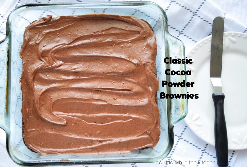 A freshly frosted cocoa powder brownies are ready to serve in a clear glass, square baking dish. The homemade chocolate frosting looks temptingly rich and smooth.