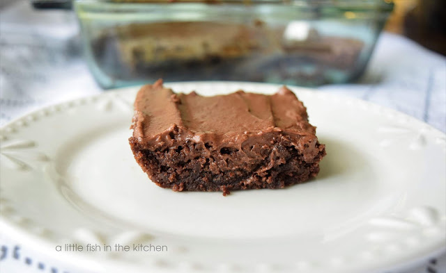 A single brownie with a luscious layer of homemade chocolate frosting is on a white serving plate and ready to be enjoyed! The dark chocolate crumb of the brownie looks moist and flavorful.