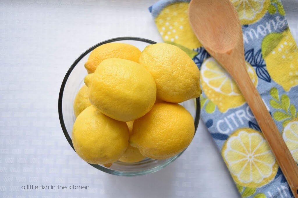A clear glass bowl is filled with bright yellow, whole fresh lemons. The bowl is on a table that is covered with a bright white tablecloth. Next to the bowl of lemons is a folded gray terrycloth tea towel with a pattern of lemons  and the word "citrus" printed on it. A wooden spoon rests on the folded novelty towel. 