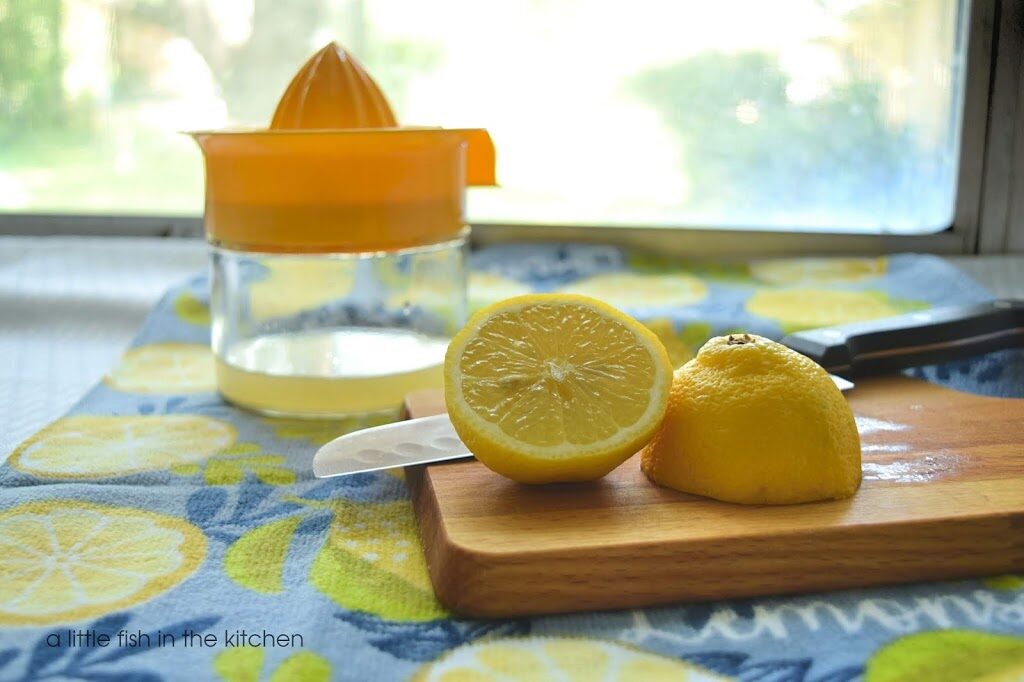 A whole fresh lemon is sliced in half on top of a small wooden cutting board. A juicer bowl partly filled with fresh lemon juice sits just behind the cutting board.  Fresh-squeezed lemon juice will be used to make a batch for tangy, refreshing homemade lemonade.