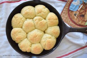 Twelve fluffy dinner rolls are inside of a black cast iron skillet. The tops of the rolls are visible and they are golden brown. The skillet sits on top of a white tea towl with a red stripes. A pot holder is off the the side of the skillet, the fabric has a picture of a horse on it. 