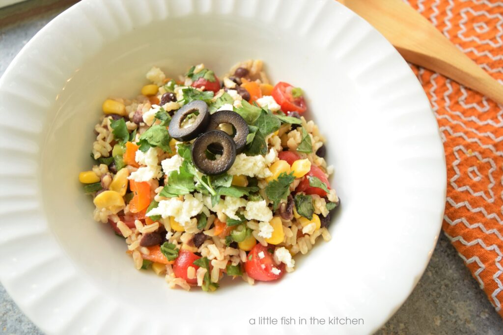 A serving of colorful, fresh Southwestern Brown rice salad is in a white bowl. Bits of fresh red and orange bell pepper, cherry tomato slices, corn kernels, black beans are visible mixed in with fluffy cooked brown rice. The spicy brown rice salad is topped with crumbed cotija cheese, chopped fresh cilantro and slices black olives. 