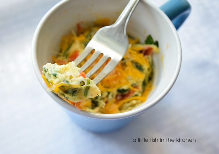 A hot omelette in a mug is studded with greens, tomatoes and topped with melty golden cheddar cheese. The eggs are in a blue ceramic mug. A fork is holding a single bite above the mug. The scrambled eggs in a mug look fluffy and tempting. 