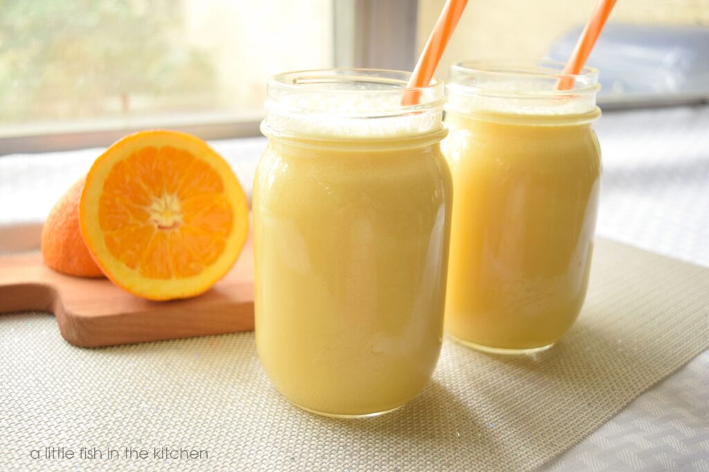 Two clear glass mason jars are filled with a frothy, creamy orange drink. The frothy tops on the drink are visible froom the sides of the glasses. Each jar has bright orange and white striped reusable straws resting on the sides. Behind the glasses of copy cat orange julius is a small wooden cutting board with a fresh sliced orange. The drinks are on a table covered with a white linen tablecloth that is near a window on a sunny day. 