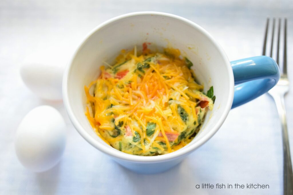 A hot omelette in a mug is studded with greens, tomatoes and topped with melty golden cheddar cheese. The eggs are in a blue ceramic mug. 