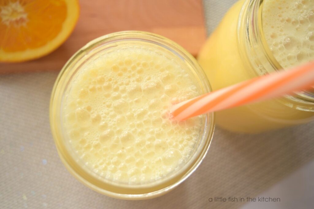The temptingly frothy top of a freshly pour glass of homemade orange julius. The blended orange drink look creamy, cold and smooth; like an orange milkshake.  The orange color of the blended citrus smoothie is pastel, but eye-catchingly vibrant.
