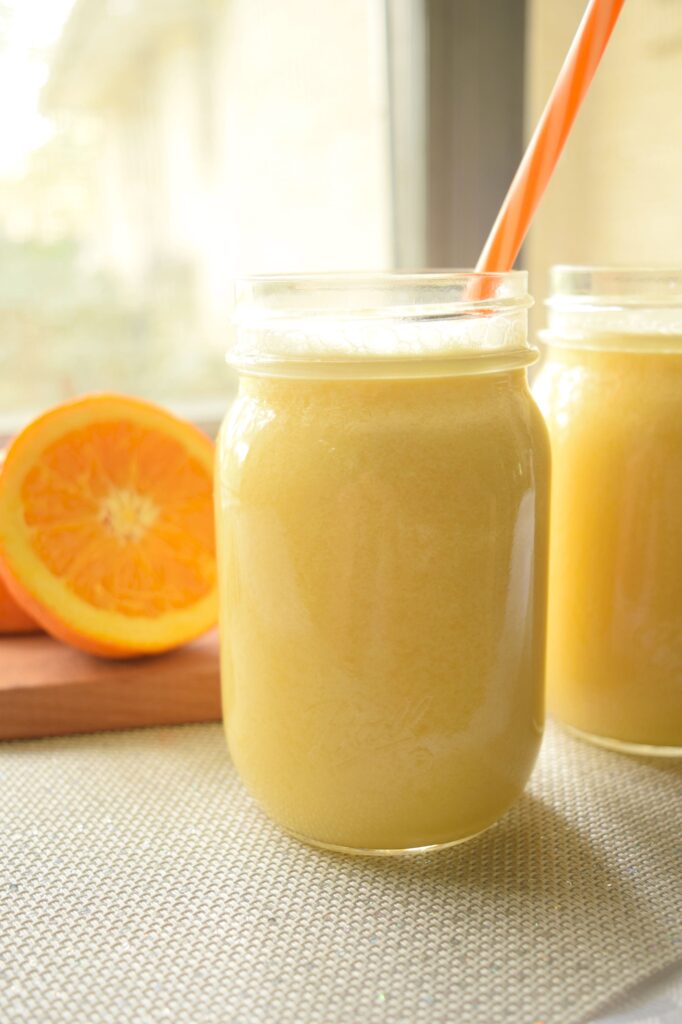 Two clear glass mason jars are filled with a frothy, creamy orange drink. The frothy tops on the drink are visible froom the sides of the glasses. Each jar has bright orange and white striped reusable straws resting on the sides. Behind the glasses of copy cat orange julius is a small wooden cutting board with a fresh sliced orange. The drinks are on a table covered with a white linen tablecloth that is near a window on a sunny day. 