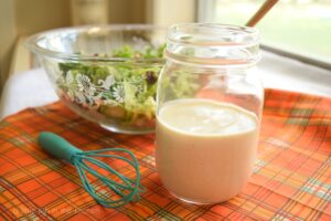 Champagne-colored creamy apple cider vinegar salad dressing is ready to serve in a clear glass mason jar. The jar of dressing sits upon a decorative piece of fabric that is plaid and features the colors orange, brown, green and gold. S small teal-colored whisk and another clear glass bowl sits nearby with a crispy, green salad in it. 