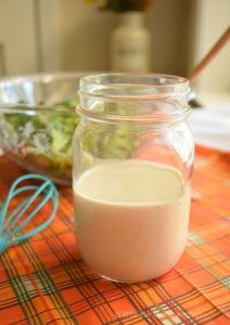 A champagne-colored salad dressing is in a clear glass mason jar and is ready to serve. A clear bowl with a green salad is slightly blurred in the background. The table is decorated with a fall-inspired piece of fabric that features the colors orange, brown, gold and green. 