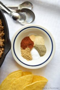 A small bowl with a blue rim has six portions of spices arranged in a circle. Chili powder, cumin, garlic powder, onion powder, salt and black pepper are visible. There is a skillet with cooked ground meat beside the bowl with the spices and crispy taco shells below the bowl.