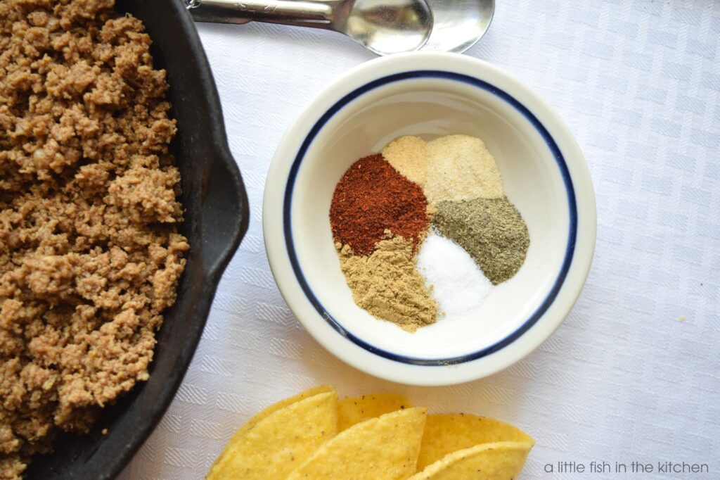 A black cast iron skillet filled with cooked ground beef is ready to serve on a table covered with a white tablecloth. Beside the skillet there is a small white round ramekin with spices arranged in a circular fashion: Chili power, onion powder, garlic powder, cumin, salt, ground black pepper will be mixed up to make easy homemade taco seasoning. 