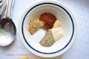 A small bowl with a blue rim has six portions of spices arranged in a circle. Chili powder, cumin, garlic powder, onion powder, salt and black pepper are visible. There are silver metal measuring spoons next to the bowl. 