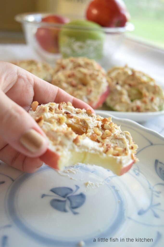 A hand holds a fresh apple slice topped with maple cream cheese. A bite has been taken and the ivory-colored apple flesh is visible. A bowl of fresh apples is slightly blurred in the background.