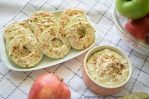 Fresh Apple Snack Cookies are ready to serve on a small white platter. A small bowl filled with flavored cream cheese is next to the plate. A small bowl full of fresh apples is next to the platter. 