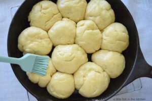 Twelve rolls of bread dough are inside of a cast iron skillet. The dough is raw and rising. A teal pastry brush is shown apply butter to the tops of the rolls to get them ready to bake. 