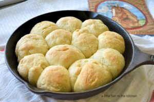 The full batch of 12 dinner rolls is shown, it looks as though they are fresh from the oven. The tops are perfectly golden brown. The skillet sits on top of a white tea towel with read stripes. A pot holder with the print of a brown horse and a blue sky is slightly blurred in the background. 