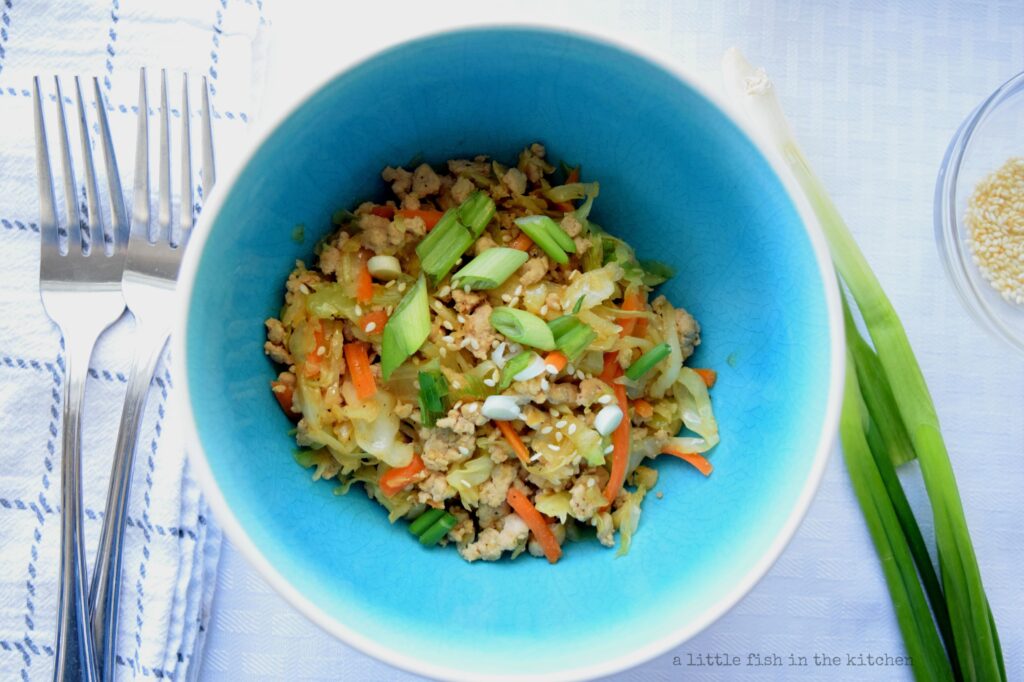 A single portion of chicken egg roll in a bowl is served in a teal ceramic bowl and is ready to eat. Ground chicken, shredded carrots and cabbage, sesame seeds and green onion are all easily visible in this savory stir fry-like dish. Two silver metal forks rest on the left side of the bowl, and a whole green onion and small bowl of sesame seeds are on the right side of the bowl. 