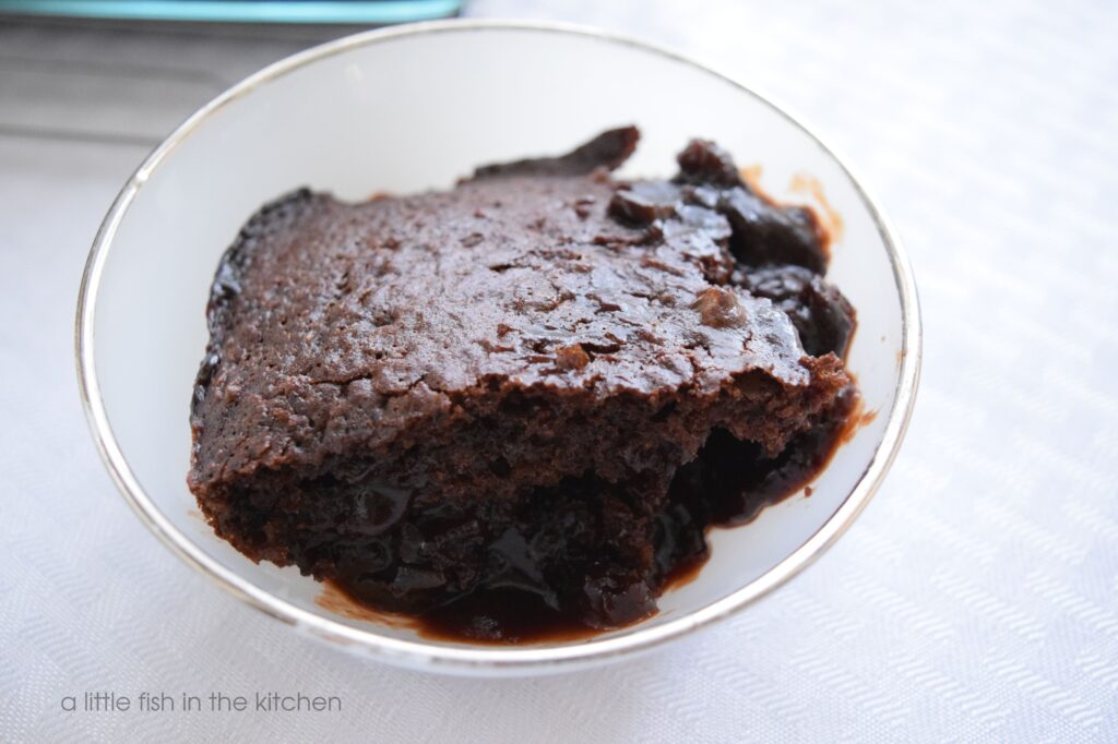 A single slice of Old-Fashioned Hot Fudge Cake rests in a small white dessert bowl. A pool of luscious chocolate sauce rests underneath the piece of moist chocolate cake. The dessert bowl is a vintage milk glass bowl with a silver rim. 