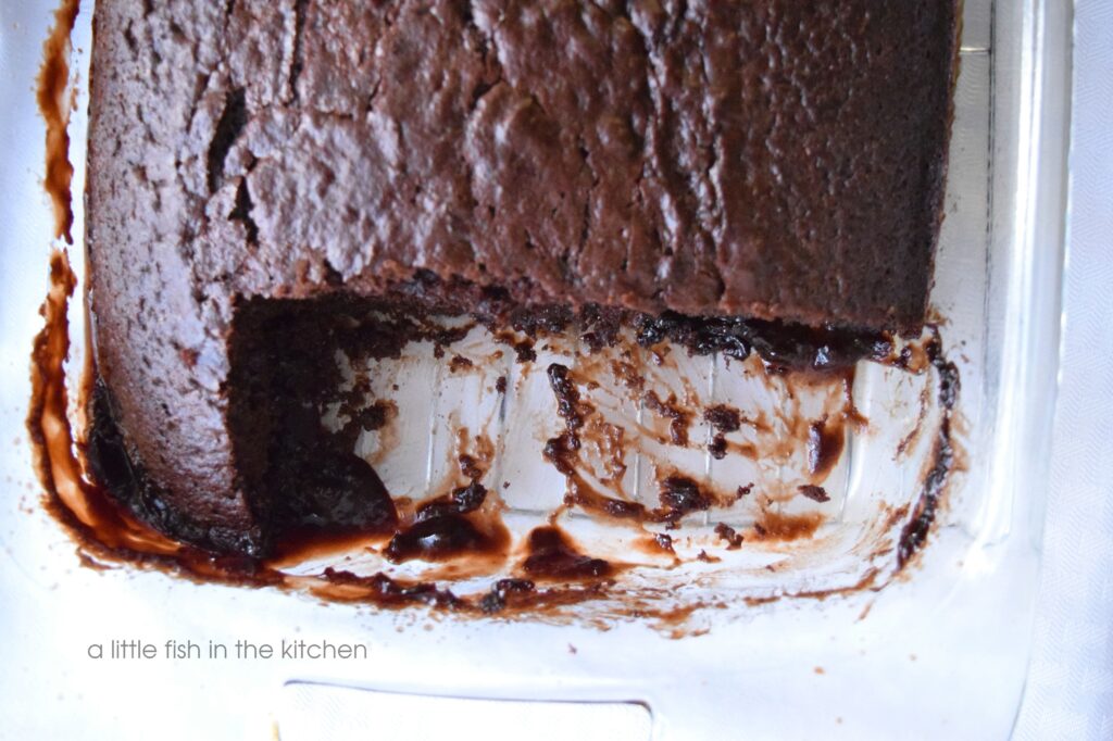 A hot fudge cake is sliced and chocolate sauce can be seen in the open spaces in the baking dish. The cake is a warm, dark brown and the crumb looks tender. The top of the rustic chocolate cake is slightly crackled. 
