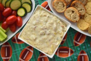 A square white bowl filled with a creamy dip sits on top of a football-themes tablecloth. There are small trays with assorted fresh veggies and sliced crackers and bread on the side. 