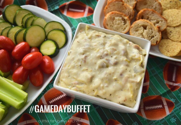Creamy chicken and artichoke Alfredo dip is served in a square, white, ceramic bowl. The creamy artichoke dip is white with bits of artichoke, ground black pepper, and cooked chicken visible in the homemade dip. Two small rectangular plates are beside the bowl, one with sliced baguette and tortilla chips, the other offers a  mix of fresh veggies, cherry tomatoes, sliced zucchini, and celery sticks, for dipping. The appetizer assortment is served on an a American football-themed tablecloth that is dark green with prints of American footballs scattered about. 