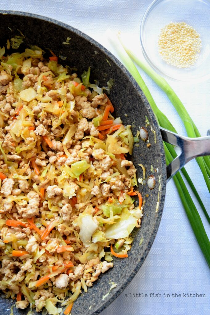Freshly cooked egg roll in a bowl is in a gray tone, non stick skillet. Bits of tender ground chicken, and shredded vegetables make up this savory mix. Two green onions and a small bowl of white sesame seeds are just beside the skillet. 