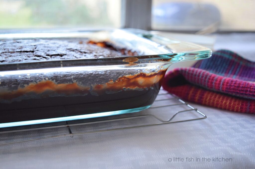A fresh baked chocolate cake is cooking on a rack. The cake is in a clear glass baking dish and decadent, bubbly chocolate sauce is seen on the side of the dish. 