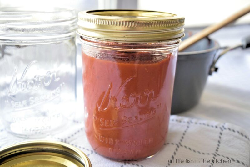 Richly red homemade pizza sauce is in a Kerr clear glass mason jar with a gold lid. The homemade pizza sauce looks thick and rich. The metal pot used to cook the sauce is slightly blurred behind the jar with a wooden spoon resting on the side. 