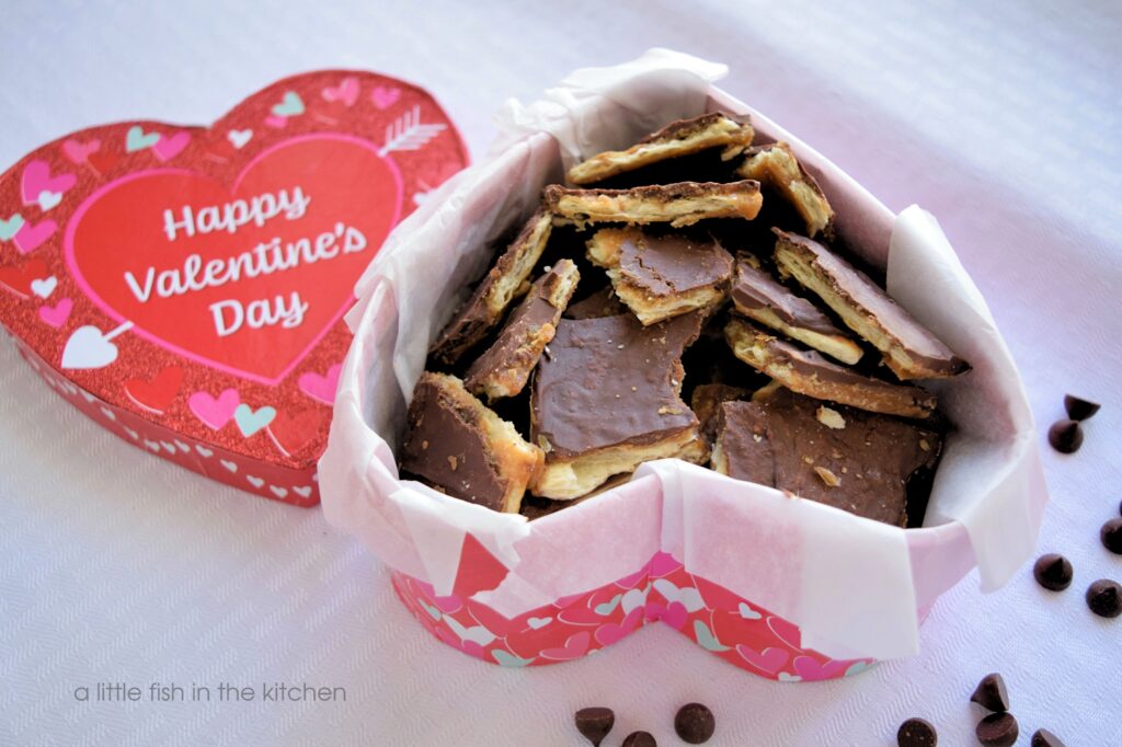 Homemade saltine cracker toffee is packed up in a heart-shaped box, a Valentine's Day gift box.  The gift box is sparkly red with pink and white hearts around the edges. The saltine cracker candy is made of visible layers of cracker, caramel and chocolate. 