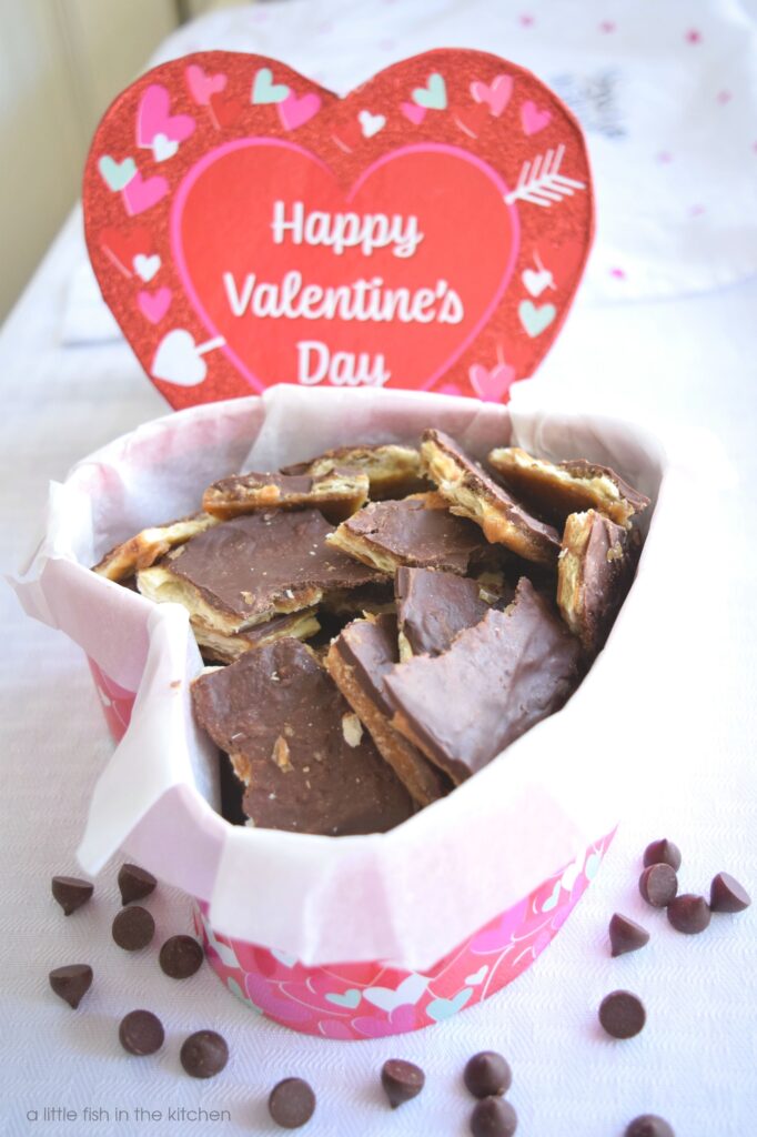 Homemade saltine cracker toffee is packed up in a heart-shaped box, a Valentine's Day gift box.  The gift box is sparkly red with pink and white hearts around the edges. The saltine cracker candy is made of visible layers of cracker, caramel and chocolate.