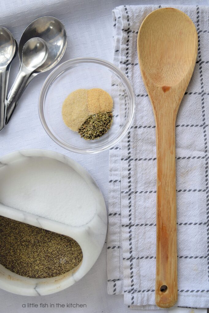 A gray and white checkered tea towel is folded in half and laid flat on a countertop. A wooden spoon is laying on the folded towel. A clear glass ramekin has measures of onion powder, garlic powder and Italian seasoning blend in it. A marble salt and pepper cellar is next to the towel; ingredients for the best homemade pizza sauce. 