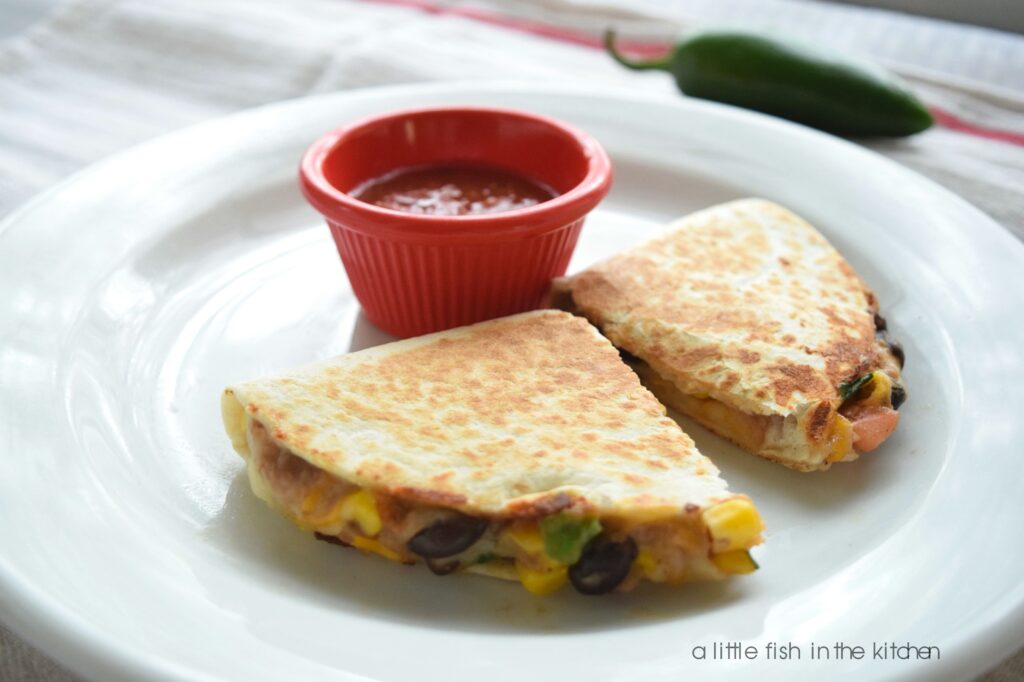 A single two-bean quesadilla is cut into two pieces and is served on a white plate with a small red ramekin filled with red salsa. From the open cut sides, kernels of corn, whole black beans, refried beans, onions and cilantro are all visible in the quesadilla filling. 