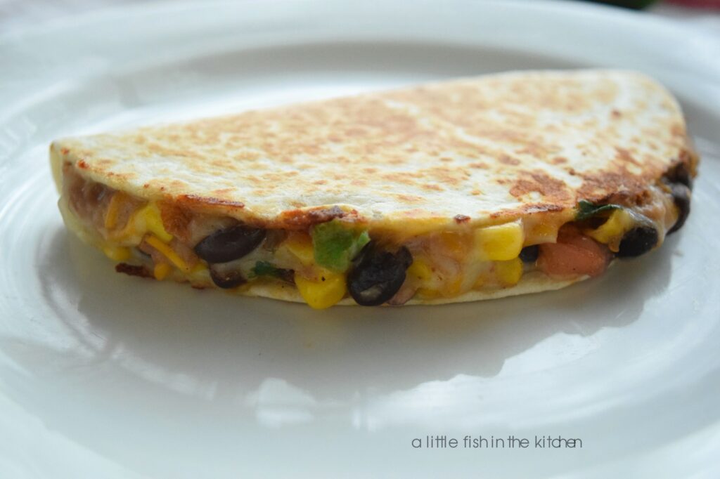 A single two-bean quesadilla is cut into two pieces and is served on a white plate with a small red ramekin filled with red salsa. From the open cut sides, kernels of corn, whole black beans, refried beans, onions and cilantro are all visible in the quesadilla filling. 